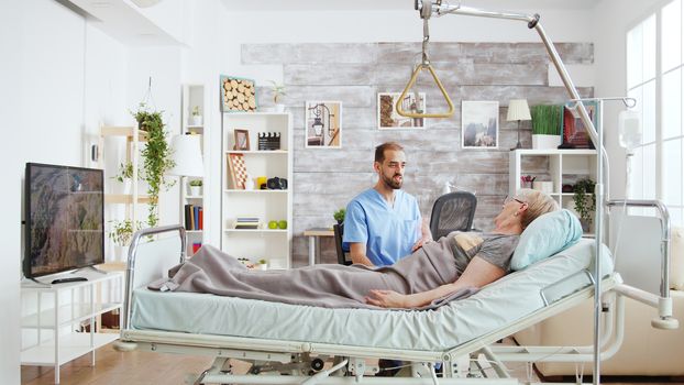 Male doctor having a conversation with ill old lady lying in hospital bed in a nursing home, medium shot.