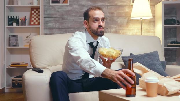 Businessman in formal wear sitting on couch eating chips while watching tv.