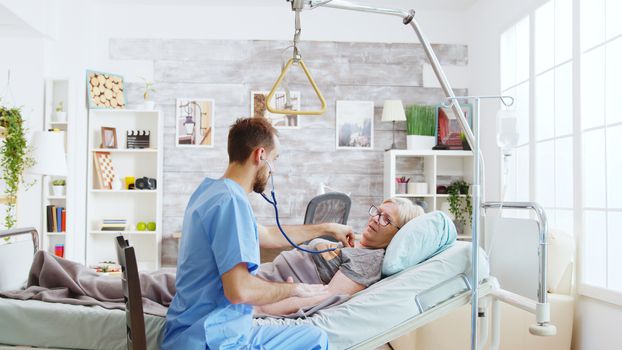 Male nurse checking the heartbeats of an sick old lady lying in hospital bed in bright and cozy nursing home