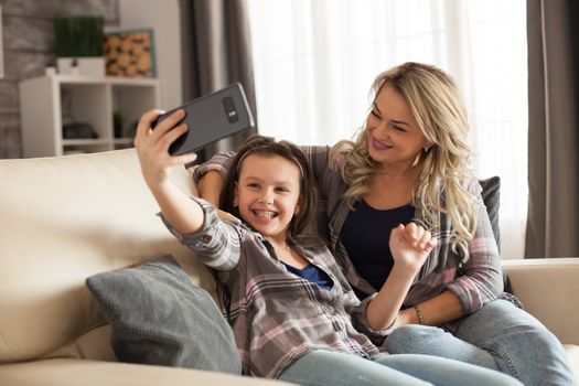 Mother and daughter bonding taking a selfie and having fun sitting on the couch in living room.
