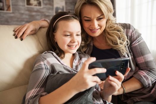 Young mother learning her little daughter with braces to use applications on smartphone sitting on the couch in living room.