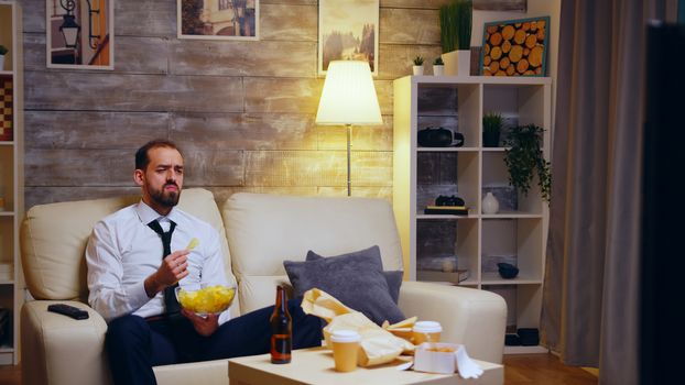 Businessman sitting on couch eating chips eating television. Zoom in shot.