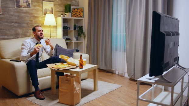 Businessman man with tie eating pizza and laughing watching tv after work sitting on couch.