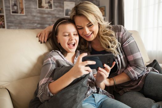 Young mother helping her little daughter with braces to use the apps from smartphone sitting on the couch in their apartment.