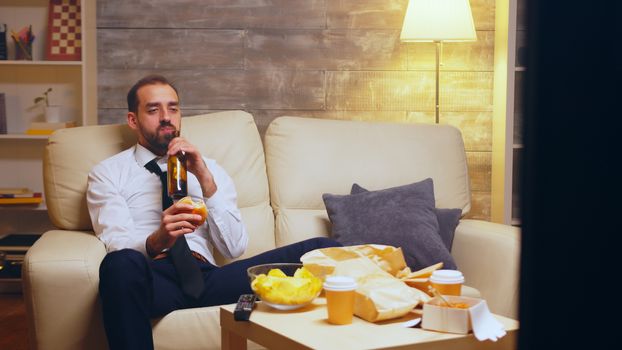 Businessman with tie sitting on couch eating a burger and talking on the phone.