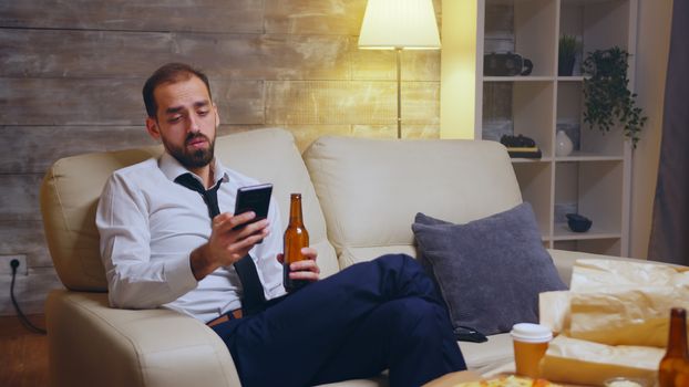 Tired businessman sitting on couch scrolling on his phone after a long day at work.