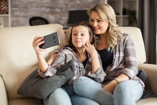 Little girl making funny faces during a video call on smartphone sitting next to her mother.