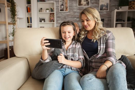 Little girl with braces and her mother on a lazy weekend using smartphone sitting on the couch.
