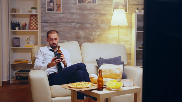 Tired businessman after long day of work browsing on phone and drinking beer. Junk food.