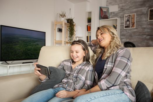 Beautiful mother and child looking at phone screen while spending time on the couch in living room.