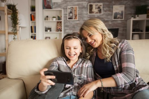 Little girl with braces and her mother having fun relaxing sitting on the couch using smartphone in living room.