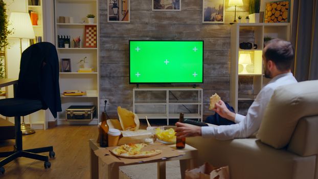 Back view of businessman enjoying his pizza while watching to a TV with Mock Up green screen