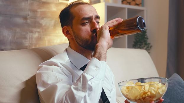 Businessman with a tie relaxing watching a comedy while eating chips and drinking beer sitting on couch.