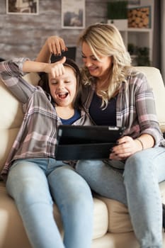 Little girl doing thumb sign during a video call on tablet sitting on the couch with her mother.