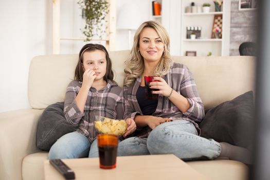 Mother and daughter watching a movie sitting on the couch on a lazy day eating chips and drinking soda.