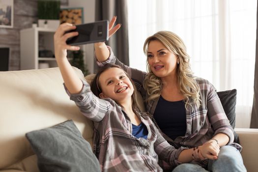 Mother and daughter in matching clothing sitting on the couch taking a funny selfie in living room.