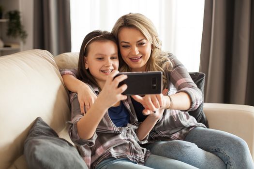 Beautiful young mother and her daughter are using a smartphone and smiling sitting on the couch in living room.