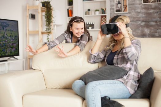 Young mother sitting on the couch in living room using virtual reality goggles. Cheerful daughter with braces.