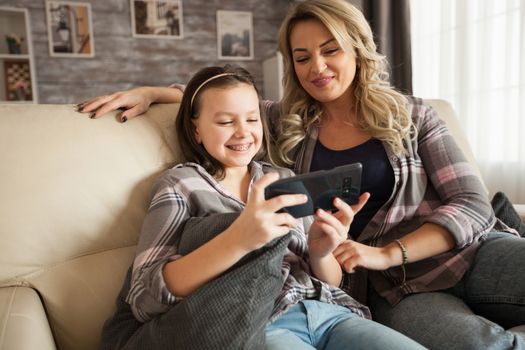Little daughter with braces and her mother sitting on the couch watching comedy on smartphone.