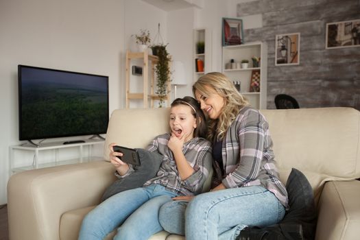 Shocked little girl white braces while using her smartphone sitting next to her mum on the couch in living room.