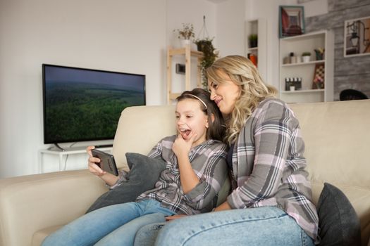 Mother and daughter making funny faces while taking a selfie sitting on the couch.