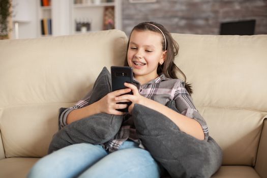 Teen girl with braces sitting on the couch in living room writing a message on smartphone .