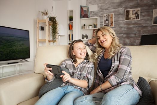 Little girl with braces smiling and pointing at phone screen sitting with her mother on the couch.