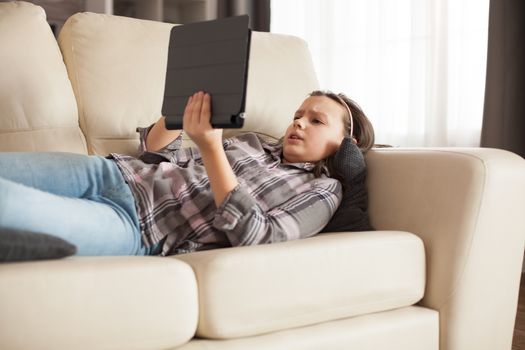 Little girl lying on sofa holding tablet. Teen watching cartoon on internet.