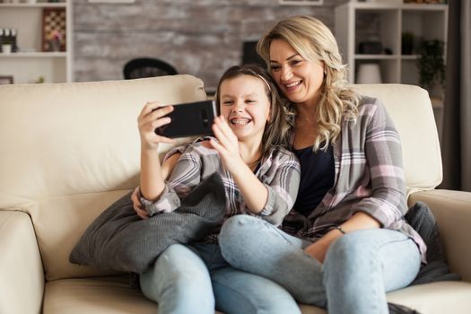 Little girl showing her braces while taking a selfie with her mother sitting on the couch in living room.