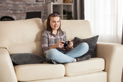 Little girl with braces sits on sofa playing video games using wireless controller.