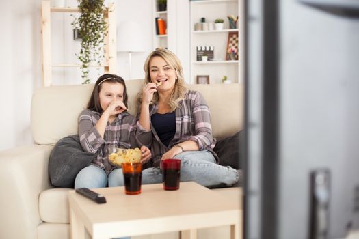 Young mother and her teenage girl watching a film on tv sitting on sofa eating chips.