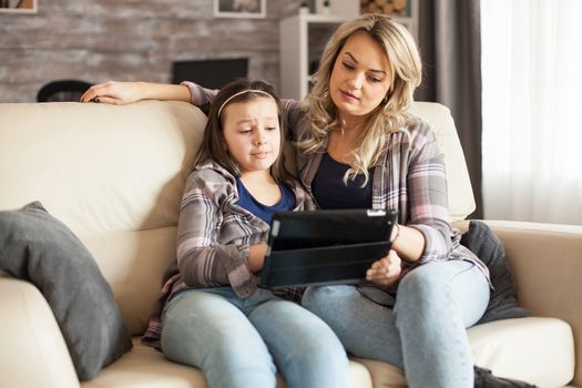 Sad little girl while watching a movie on tablet sitting on the couch with her mother.