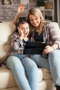 Cheerful little girl during a video call on tablet with her grandparents sitting on the couch with her mother.