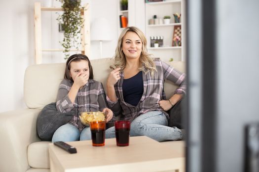 Young mother and her little girl watching movies on big television in living room. eating chips and drinking soda.