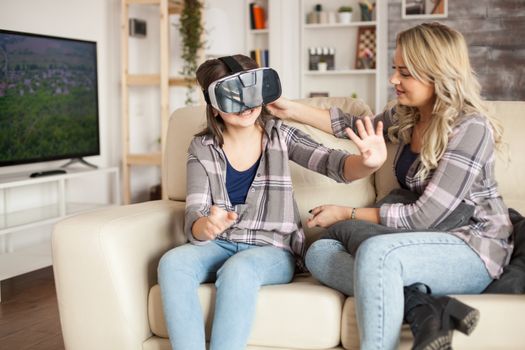 Little girl playing video games using virtual reality goggles sitting on the couch next to her mother.