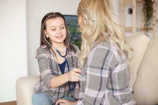 Little daughter listening mother's heart sitting on the sofa using a stethoscope.