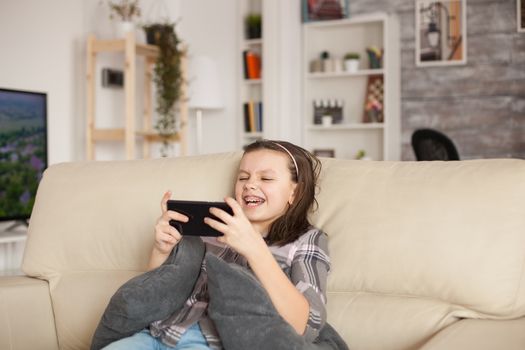 Happy little girl playing games on smartphone sitting on the couch in living room.