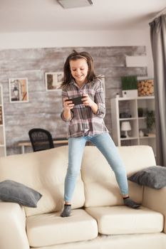 Excited little girl using her smartphone and jumping on the sofa.