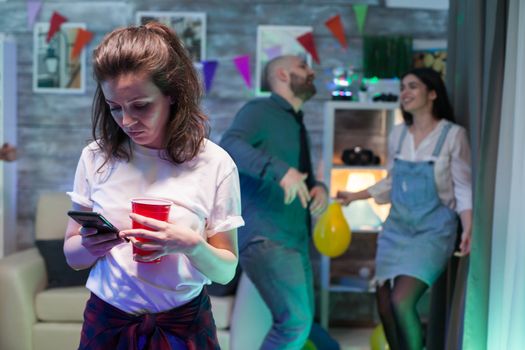 Young woman using her smartphone while her friends are celebrating and dancing in the background.