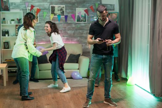 Bored young man using his smartphone at a party while his friends ar dancing.