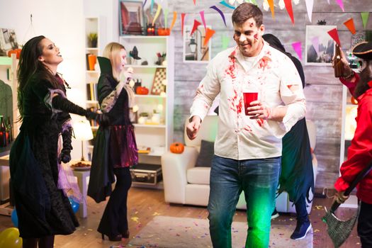 Man dressed up like a zombie drinking beer while celebrating halloween with his friends.