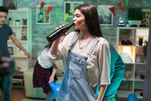 Cheerful young woman at a party with her friends taking a sip of beer. Young people dancing.