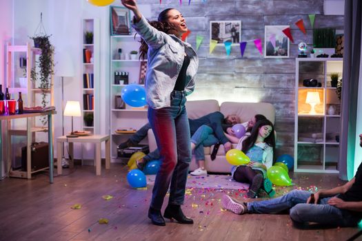 Cheerful young woman holding a balloon while dancing at the party while her friends are lying on the floor.