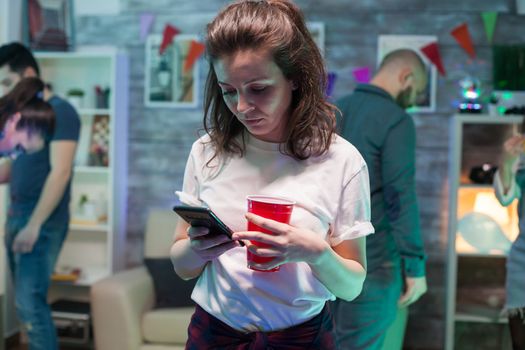 Beautiful young woman using her smartphone at her friend's party and holding a beer cup.