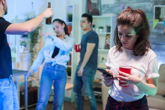 Shy young woman using her smartphone while her friends are dancing.