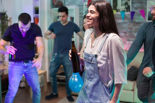 Cheerful young woman smiling and holding a beer bottle at a friend party.