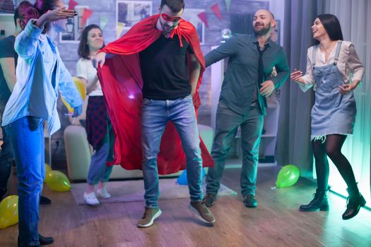 Young man wearing a superhero costume in a room with balloons dancing with his friends.