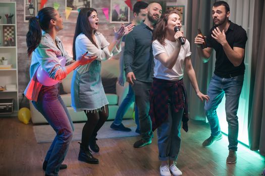 Cheerful young woman singing on microphone while her friends are dancing at the party.