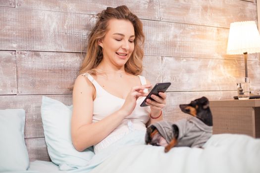 Beautiful woman in pajamas smiling while using her phone at night in bed. Little dog.