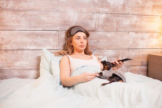 Beautiful young woman with eye cover mask watching tv in bed with her little dog.